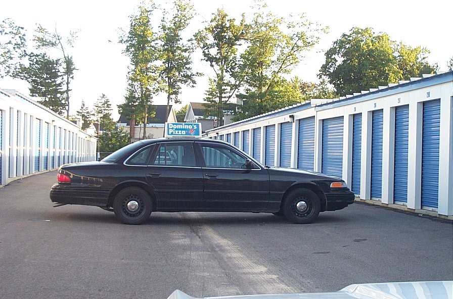Side view of a black 2001 Ford Crown Victoria with a Domino's Pizza sign on the roof, parked in front of storage units