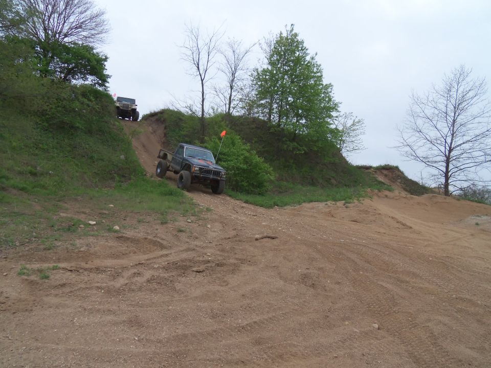 Two off-road trucks driving up a steep dirt hill with trees and overcast sky