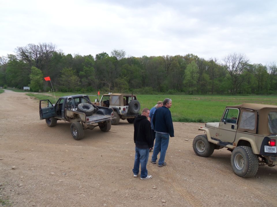 Several off-road vehicles and four people stand on a dirt road near a grassy field and trees