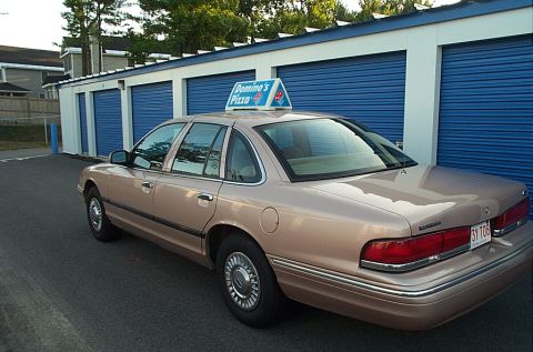 2001 Ford Crown Victoria - A tan 2001 Ford Crown Victoria sedan, equipped with a Domino's Pizza sign, is parked outside a row of blue storage unit doors.