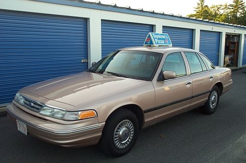 2001 Ford Crown Victoria - A tan 2001 Ford Crown Victoria, likely used for pizza delivery, is parked in front of blue storage unit doors.