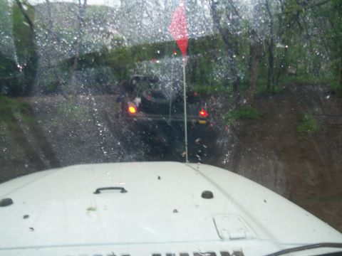 1989 Ford Bronco - Two SUVs navigate a muddy off-road trail, viewed from the hood of a white Jeep.