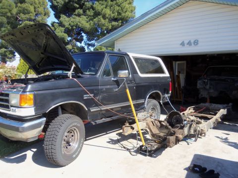 1968 Ford Bronco - A black Ford Bronco is shown with its hood open and the chassis removed, parked outside a garage where another vehicle frame is visible.