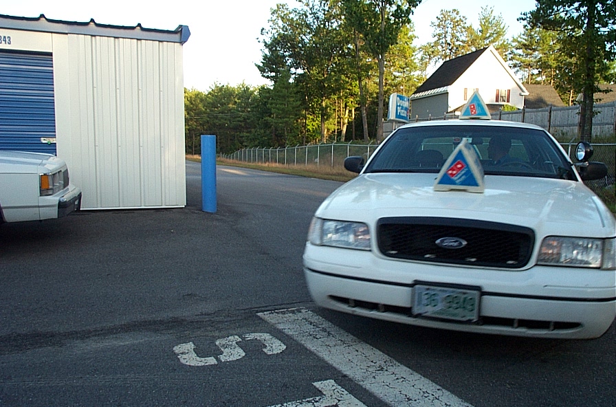 White 2001 Ford Crown Victoria with Domino's pizza signs on the roof and hood, parked next to a storage unit