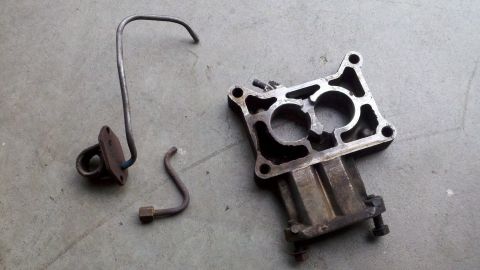 1978 Ford Bronco - Two separate automotive parts, a bent metal fuel line and a metal carburetor base, are shown on a concrete surface.