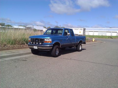 1992 Ford F-150 - A blue 1992 Ford F-150 pickup truck is parked on the side of the road under a partly cloudy sky.