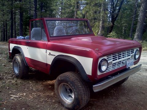 1968 Ford Bronco - This two-tone red and white 1968 Ford Bronco is shown in a wooded area, its iconic grille and chrome bumper.
