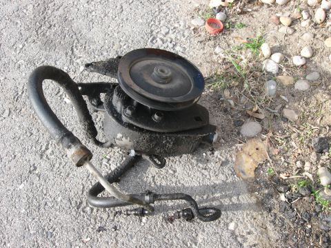 1981 Ford Bronco - A greasy power steering pump with its pulley and hoses lies on a gravelly surface.
