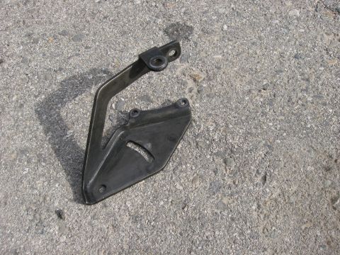 1981 Ford Bronco - A close-up view of a vehicle part, a metal bracket connected to a plastic shield, resting on a rough asphalt surface.