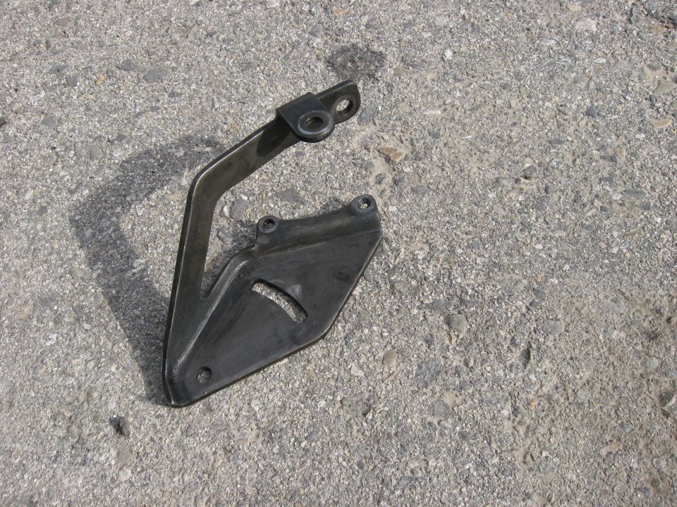Metal bracket attached to a black plastic shield with a curved cutout, lying on asphalt