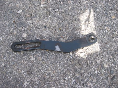 1981 Ford Bronco - A dark metal bracket with a slot and hole is pictured on an asphalt surface.