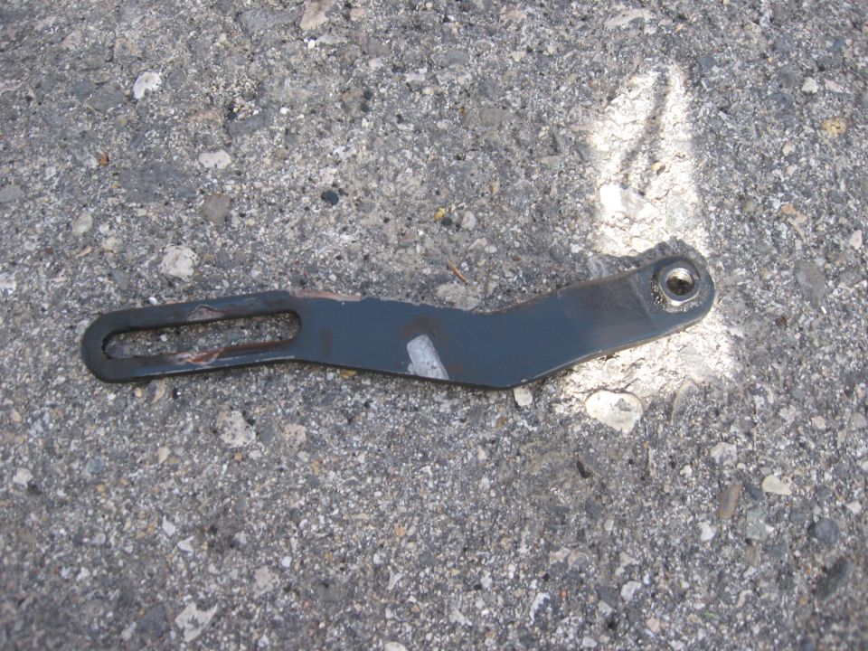 Dark metal bracket with an elongated slot and a circular hole, lying on asphalt