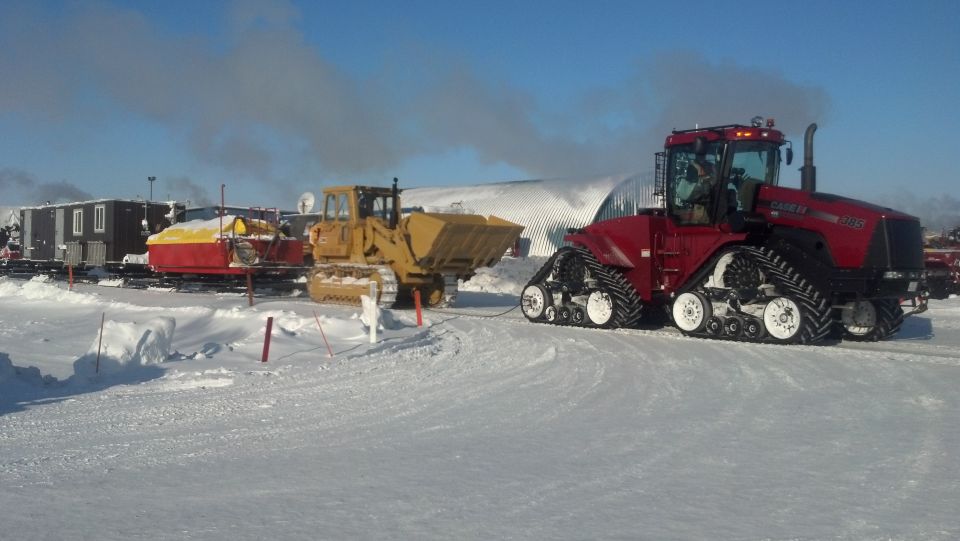 Red tracked tractor and yellow bulldozer parked in a snowy area with buildings in the background