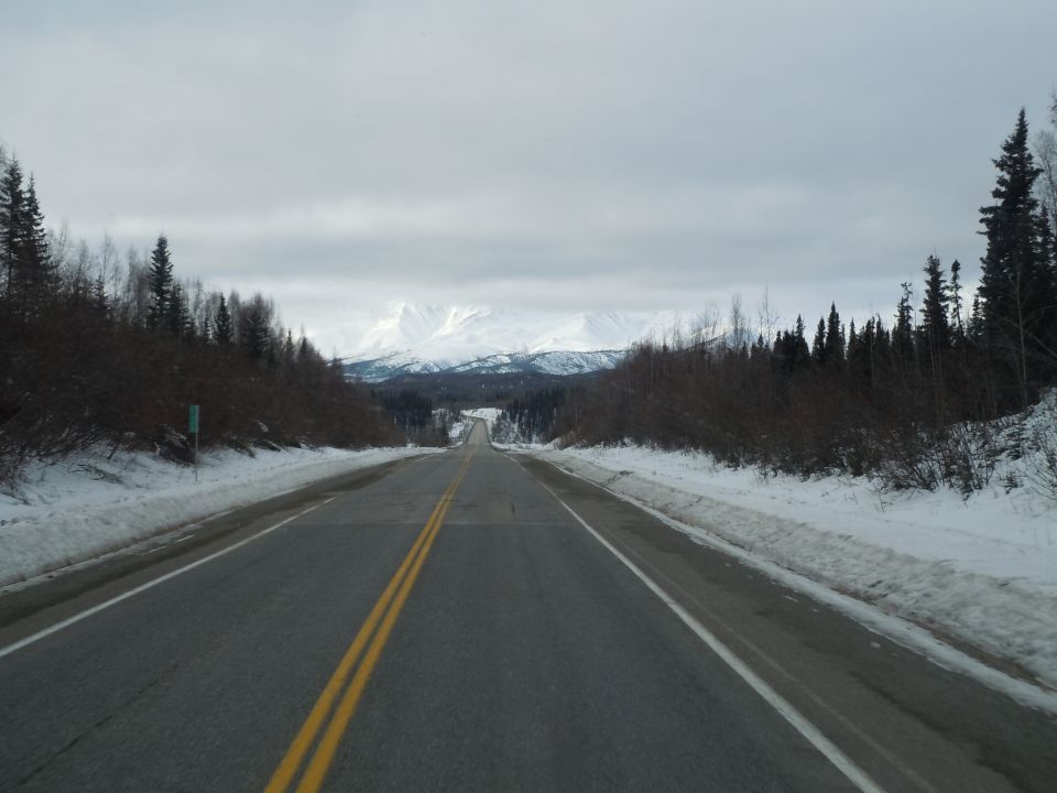 Snowy road leading to mountains under an overcast sky