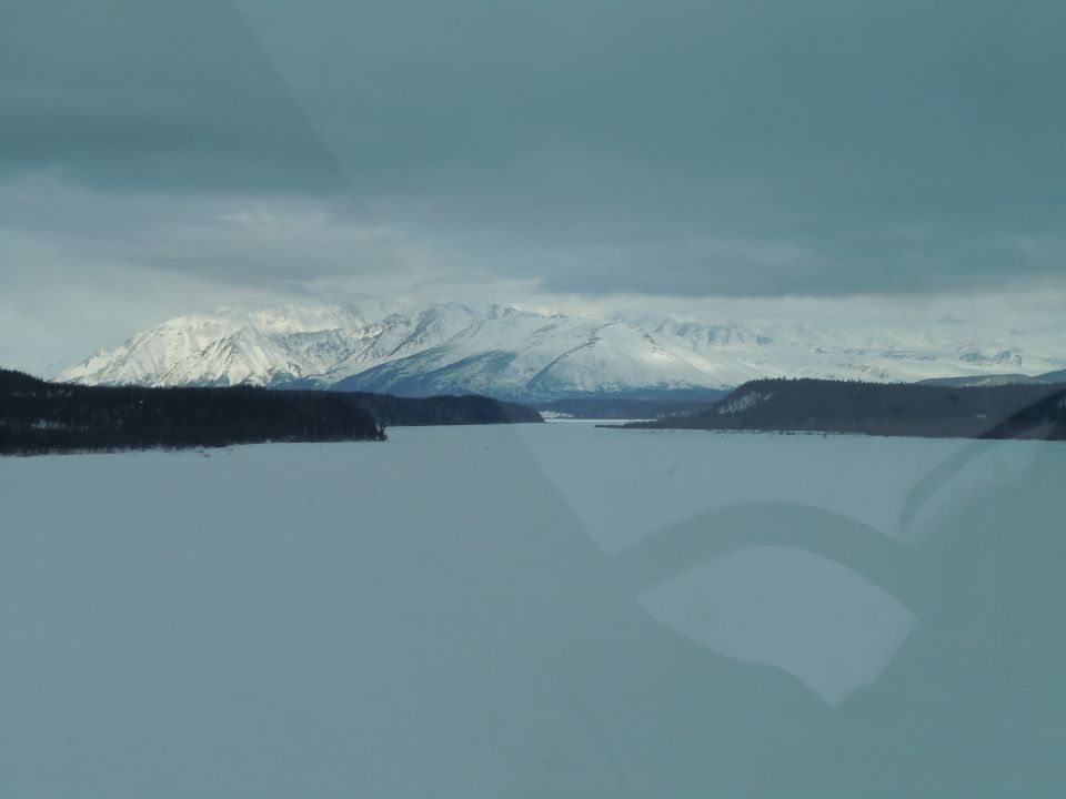 Snowy mountains overlook a frozen lake under a cloudy sky. Watermark reads SUPERMOTORS