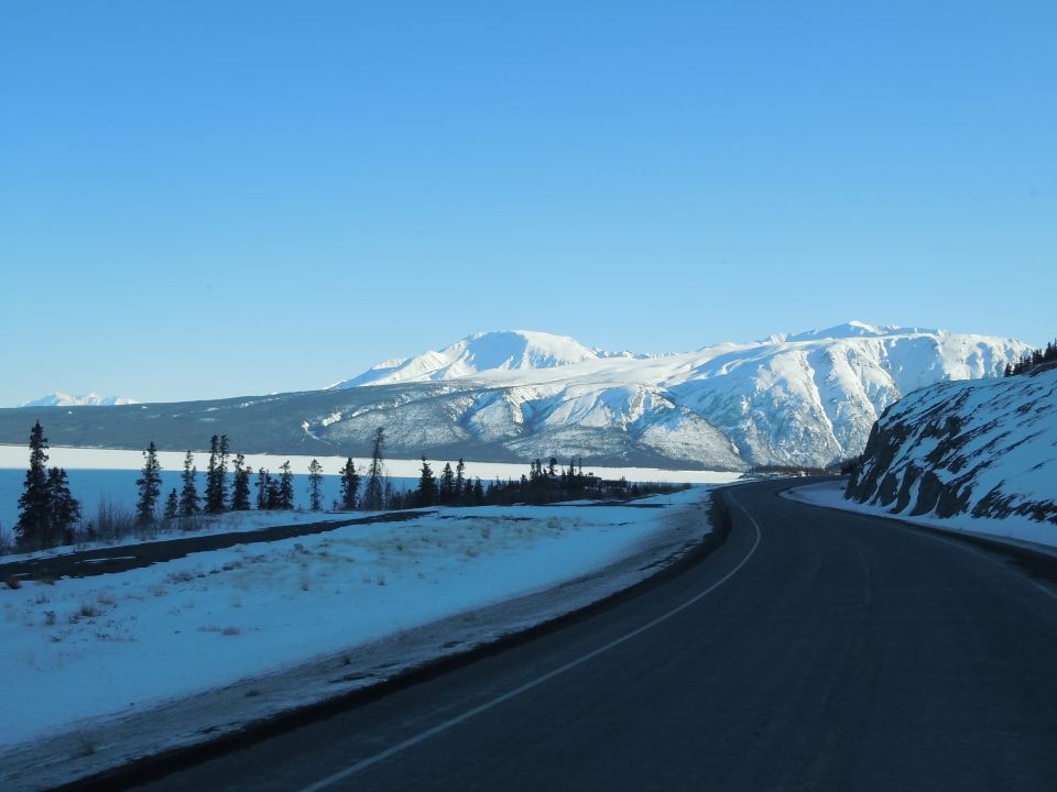 road curves through snowy mountains with a frozen lake on the left