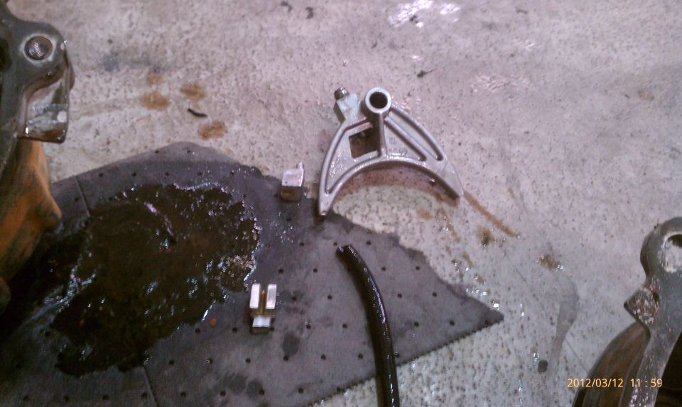 Metal automotive part with a curved arm and a hole, lying on a shop floor next to a dark fluid spill