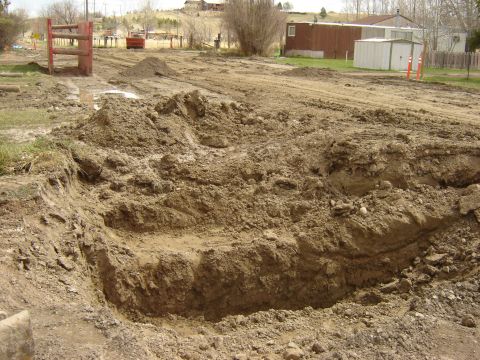 1984 Ford Mustang - A construction site features a deep excavation in the foreground, with a wooden barrier and a red dumpster visible in the distance.