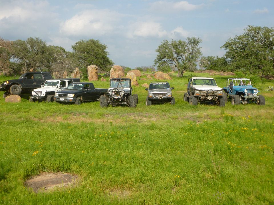 Several modified off-road vehicles parked in a grassy field with large rocks and trees in the background