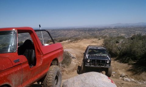 1978 Ford Bronco - Two off-road vehicles navigate a dusty trail with a scenic overlook in the background.