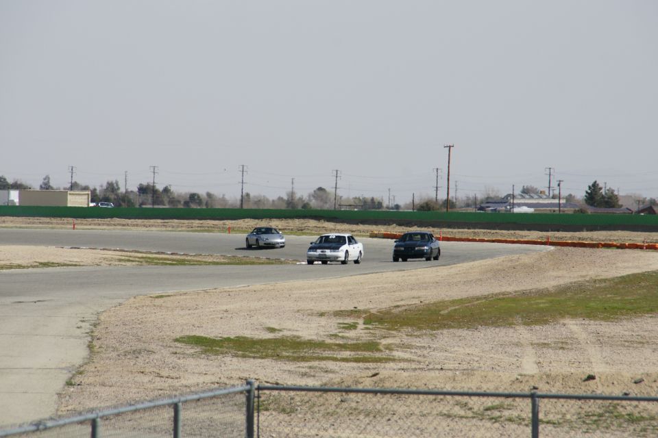 Three sedans on a race track, one white with black hood in the lead