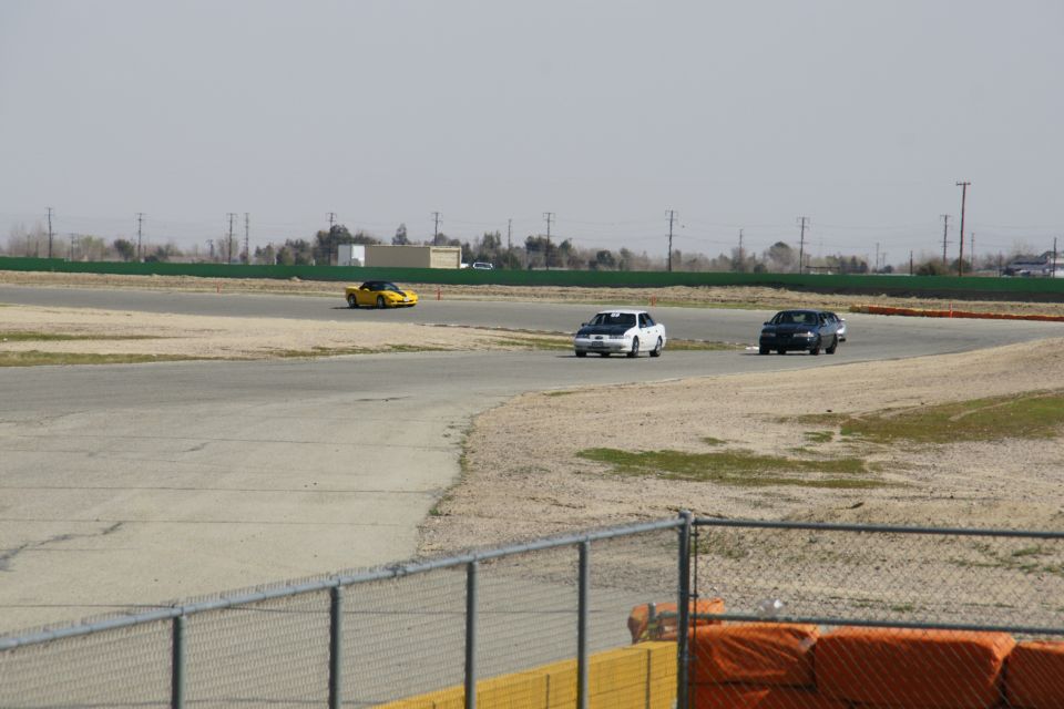 Cars on a race track, one yellow sports car in distance, white and dark sedans closer