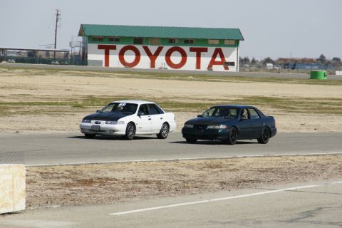 1995 Ford Taurus - Two sedans, one a 1995 Ford Taurus, race on a track with a Toyota building visible.