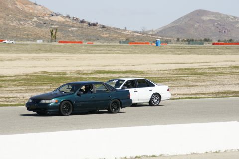 1995 Ford Taurus - Two sedans, a green 1995 Ford Taurus and a white car, race on a track with hills in the background.
