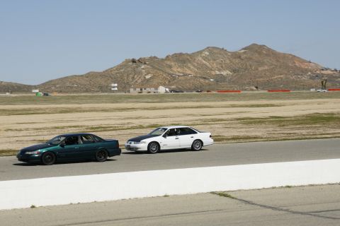 1995 Ford Taurus - Two sedans drive on a paved track with a barren, mountainous landscape under a clear sky.