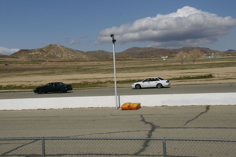 Two sedans race on a track with dry hills and a cloudy sky in the background