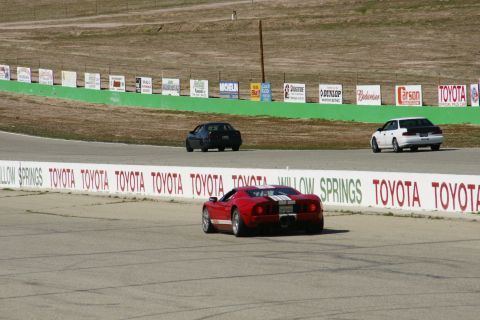 1995 Ford Taurus - A red sports car with white racing stripes navigates a turn on a race track, with other vehicles in the background.