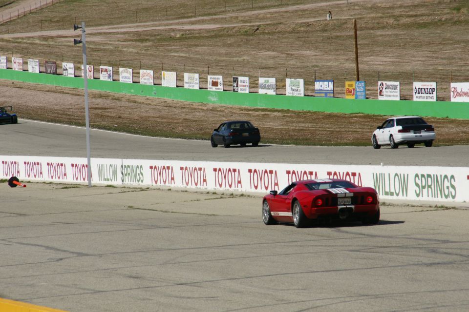 Race track scene with a red sports car, a white sedan, and a dark sedan on the track. Toyota and Willow Springs signage visible
