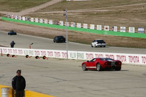 1995 Ford Taurus - A red sports car with white racing stripes drives on a track, with other vehicles and spectators visible.