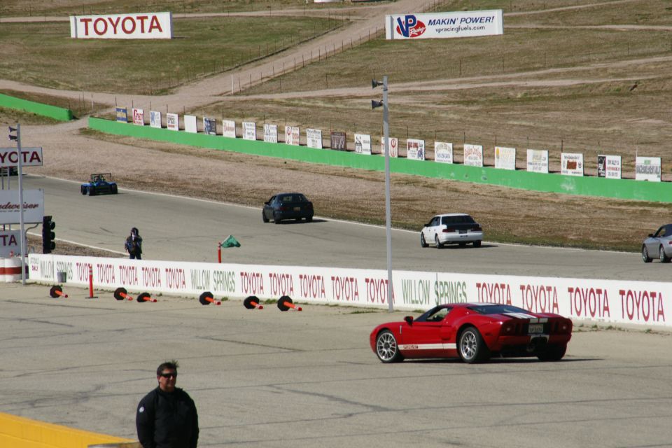 Red sports car with white racing stripes on a race track, other cars and track signage visible