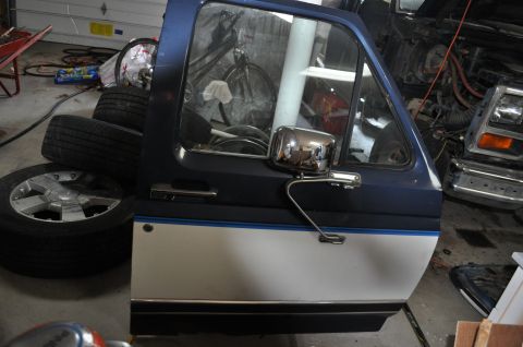1986 Ford Bronco - A blue and white Ford Bronco door is visible, with tires stacked next to it and another vehicle in the background.