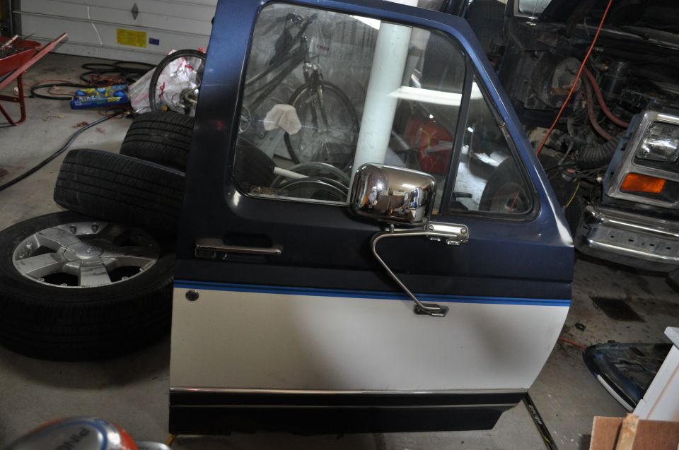Blue and white Ford Bronco door with chrome mirror, stacked tires nearby, another vehicle in background