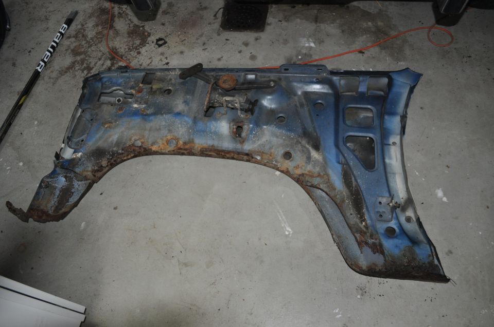 Rusted blue fender assembly with some mounting brackets attached, lying on a concrete floor. A hockey stick is visible to the left