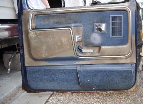 1986 Ford Bronco - The interior door panel of a vehicle shows signs of wear and tear, with a blue fabric lower section and a textured brown upper section.