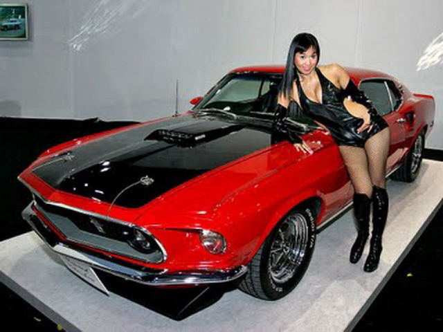 Red 1969 Ford Mustang Mach 1 with black hood scoop and stripes, woman in black outfit posing beside it