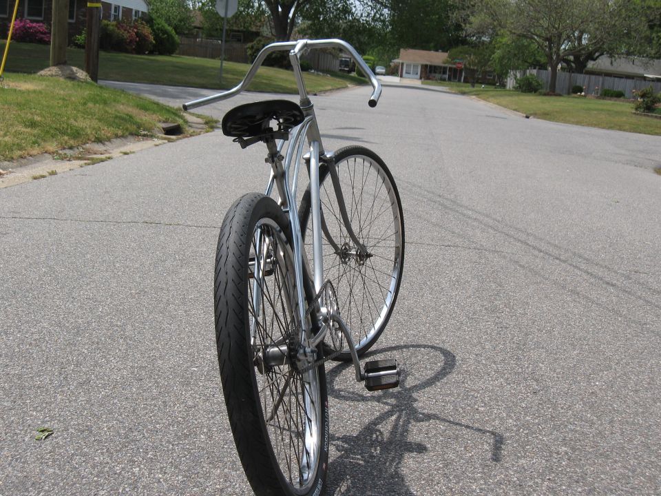 Rear view of a vintage cruiser bicycle with wide handlebars and a black seat, parked on an asphalt road