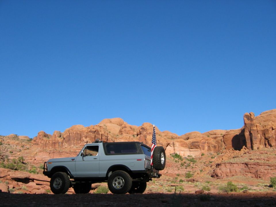 Light blue 1978 Ford Bronco with spare tire and American flag parked in desert landscape