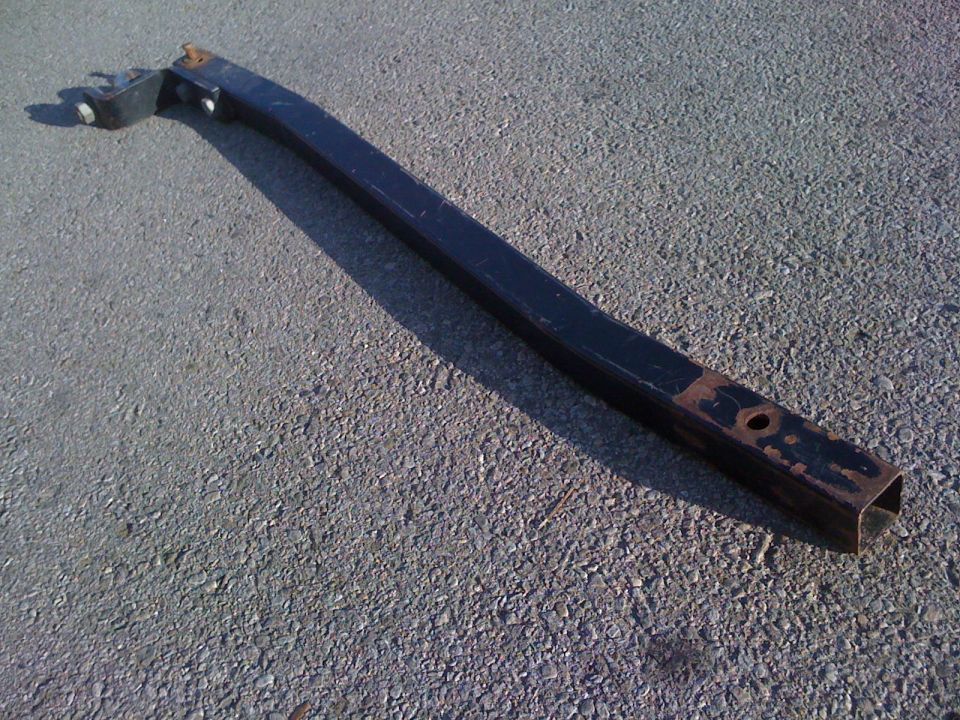 Black square-tube steel frame rail with end bracket and rusted mounting hole, lying on asphalt