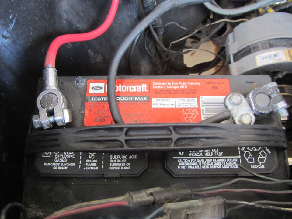 Close-up of a car battery with red and gray cables attached. The battery label says Motorcraft Tested Tough Max