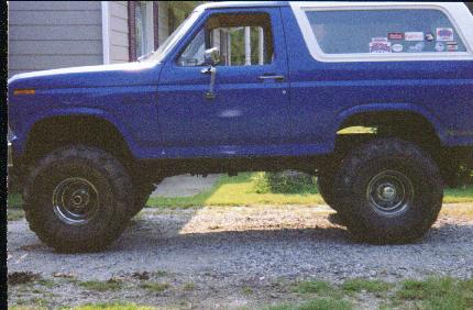 1985 Ford Bronco - This lifted blue Ford Bronco sits on a gravel surface, showing its side profile and large tires.