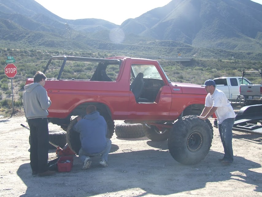 Red 1978 Ford Bronco with two people working on the front tire and one person on the rear tire. A white pickup truck is visible in the background