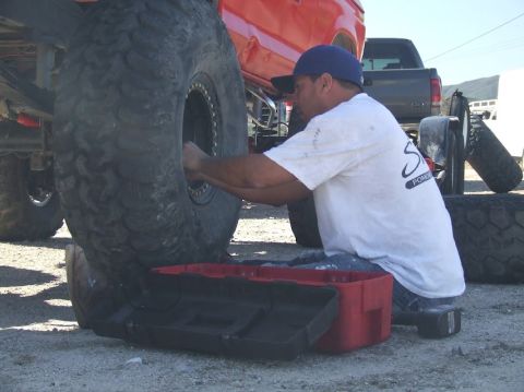 1978 Ford Bronco - A man kneels to work on a massive tire, with a red truck and another vehicle visible in the background.