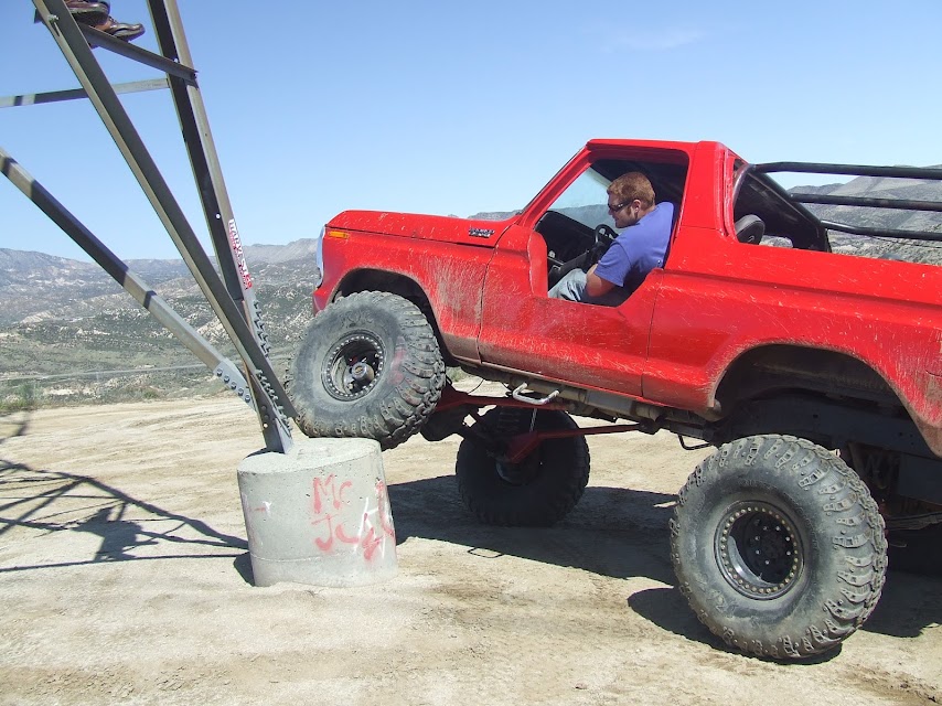 Red 1978 Ford Bronco with large off-road tires is climbing over a concrete pillar
