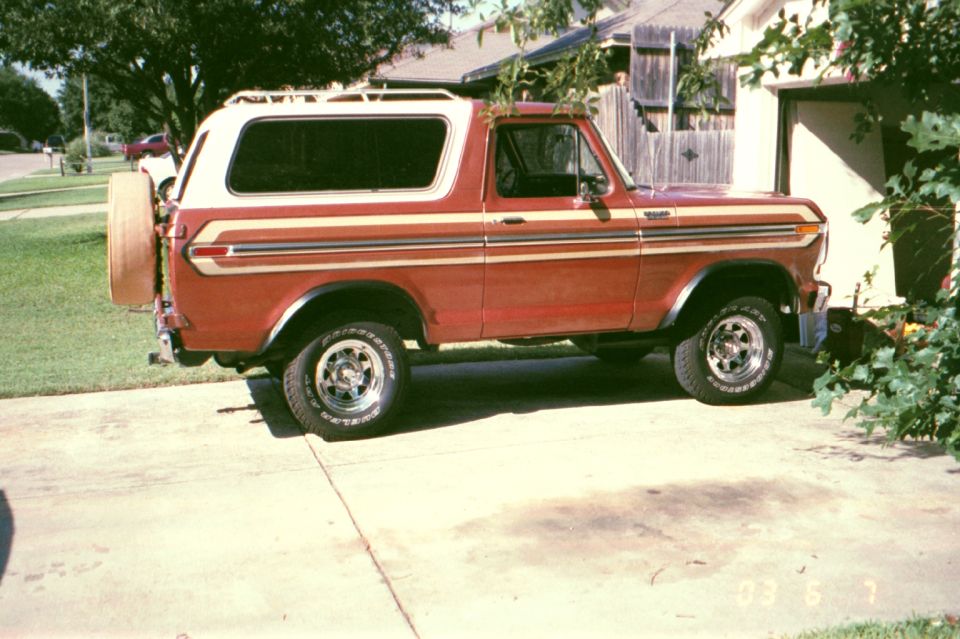 Red 1979 Ford Bronco with white roof, side view, parked in a residential driveway with rear-mounted spare tire.