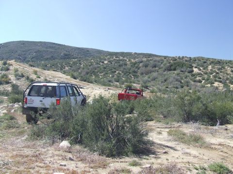 1978 Ford Bronco - Two off-road vehicles navigate a dusty trail through a scrub-covered hillside under a clear blue sky.