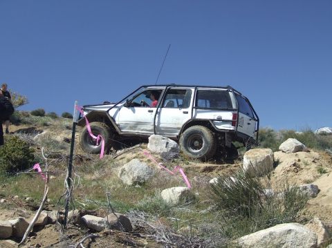 1978 Ford Bronco - White SUV with roll cage navigating a rocky off-road course under a clear blue sky.